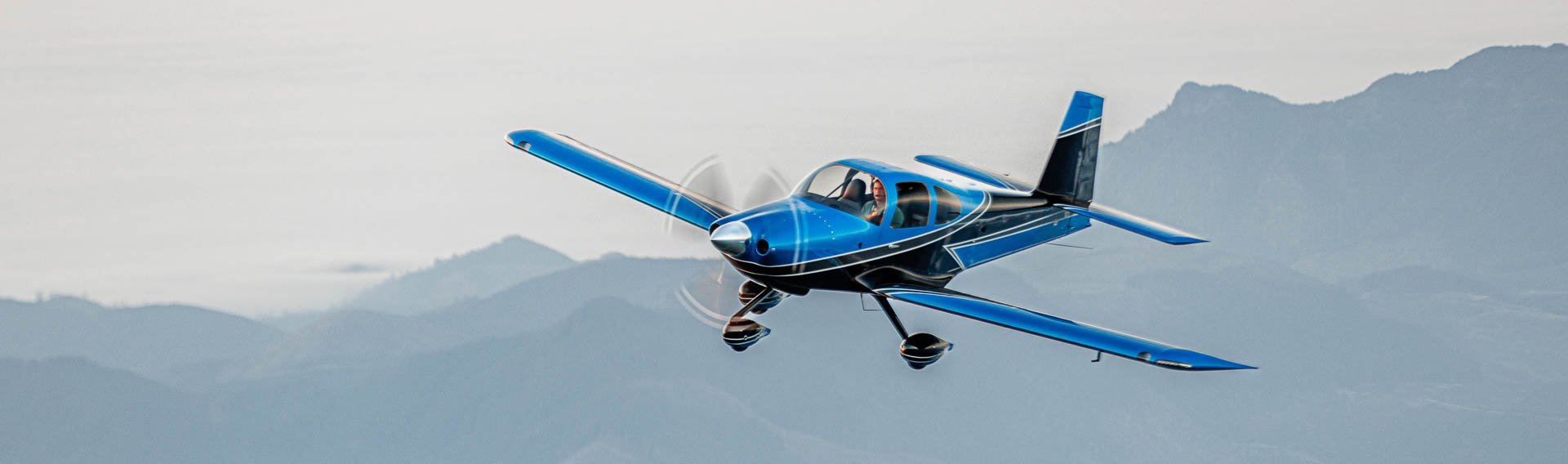 Van’s Aircraft RV-10 four-seat, single-engine experimental airplane flying above a cloud layer with mountains in the background.